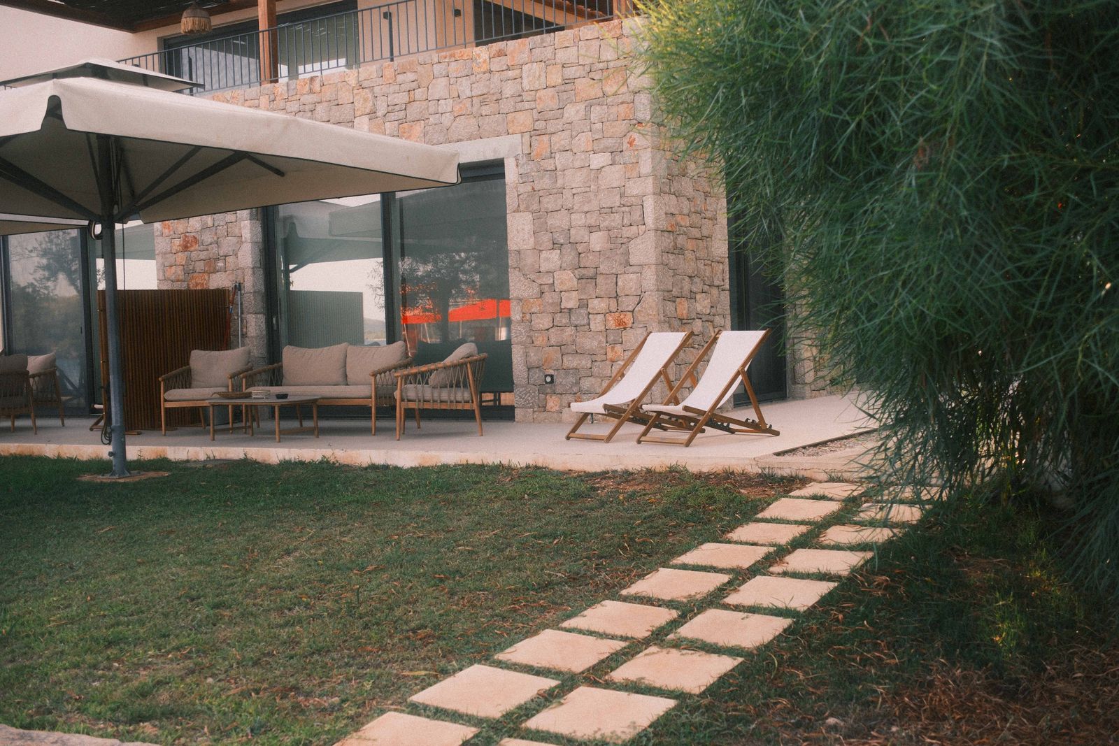 Relax in style on a beautiful stone patio with outdoor furniture, greenery, and loungers, perfect for a summer retreat.