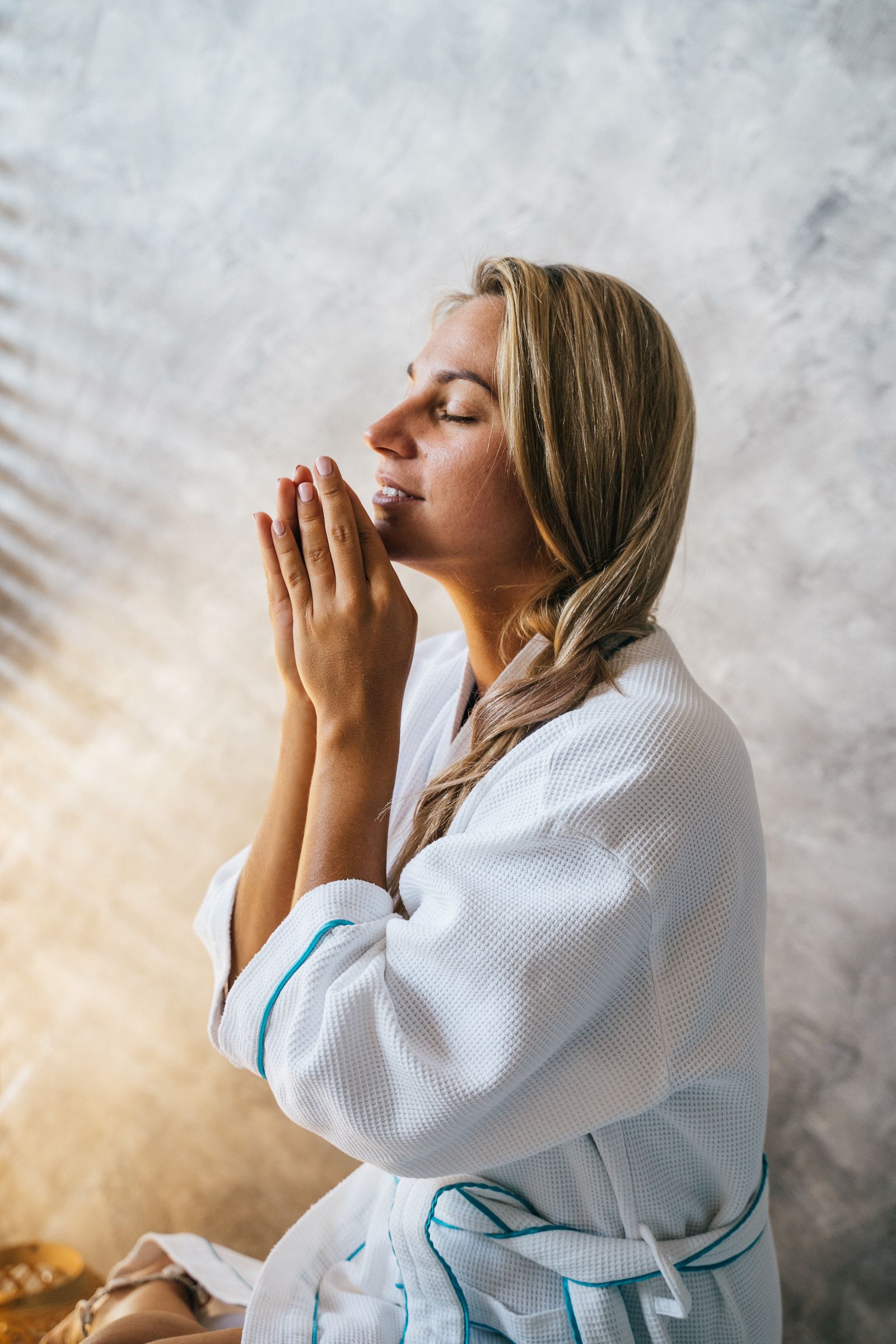 Serene Caucasian woman in a bathrobe with hands together, eyes closed, enjoying a spa treatment indoors.